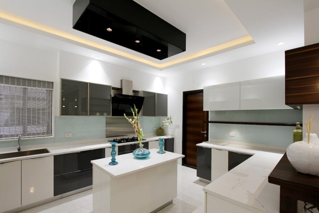 Contemporary kitchen interior design demonstrating layered residential lighting, including cool white LED recessed downlights, warm white under-cabinet task lighting, and a decorative black ceiling light feature over a central white island