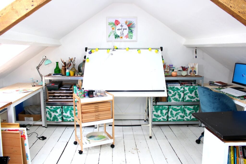 A brightly lit loft art studio and home office under sloped ceilings with exposed beams. The workspace utilizes high-output cool white task lighting from a clamp-on desk lamp over a large central drawing board and an adjustable mint-green desk lamp, making it ideal for precision craft work, painting, and computer tasks.