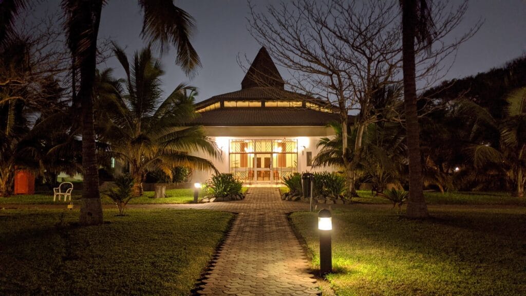 A beautifully illuminated tropical garden path at night, featuring low-level black bollard lights casting a warm white glow along a brick walkway. The path leads to a prominent pavilion building with bright interior lighting, surrounded by uplit palm trees and landscaped greenery